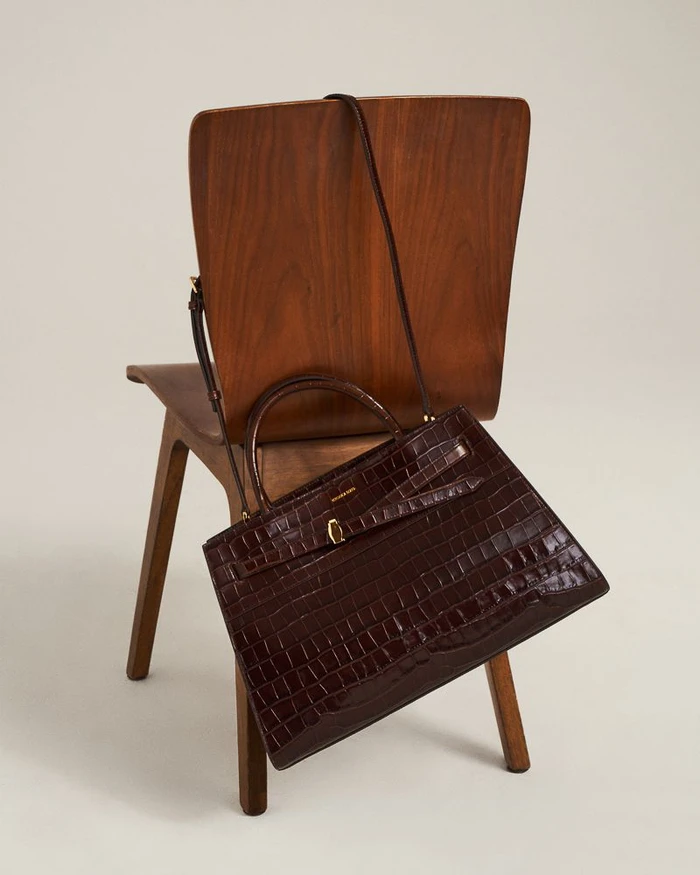 Dark brown croc-embossed leather tote bag resting on a mid-century wooden chair against a plain studio background.