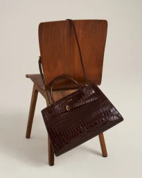 Dark brown croc-embossed leather tote bag resting on a mid-century wooden chair against a plain studio background.