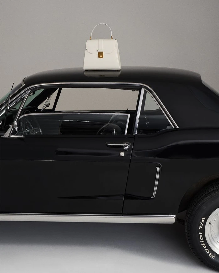 A white structured handbag with a top handle resting on the roof of a vintage black muscle car in a studio setting.