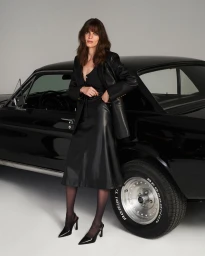 Model wearing a black leather blazer and midi skirt posing against a classic black car on a studio background.