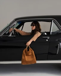 Lola Mcdonnell wearing sunglasses in a classic black car holding a brown suede handbag outside the window.