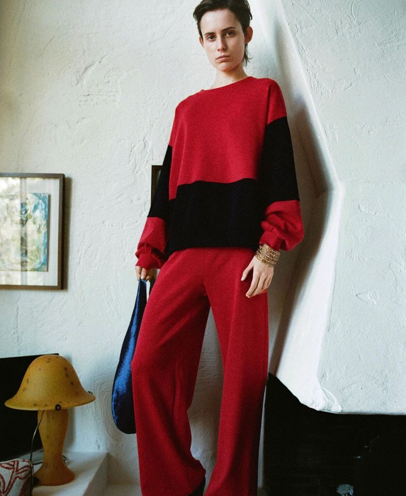 A young adult woman in a red and black sweater and red pants poses in an indoor setting with a lamp and painting visible.