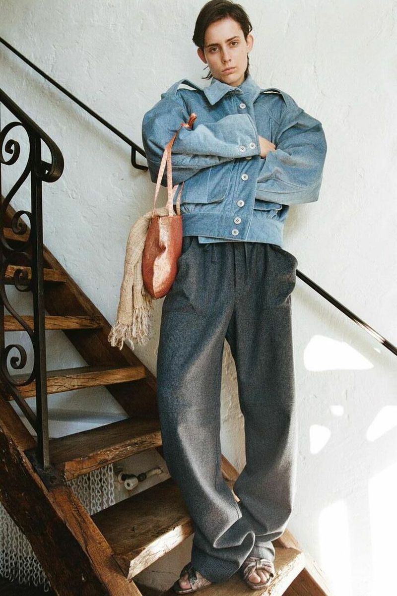 Woman in a blue jacket and grey trousers poses on wooden stairs indoors with a unique handbag and scarf, showcasing fashion and style.
