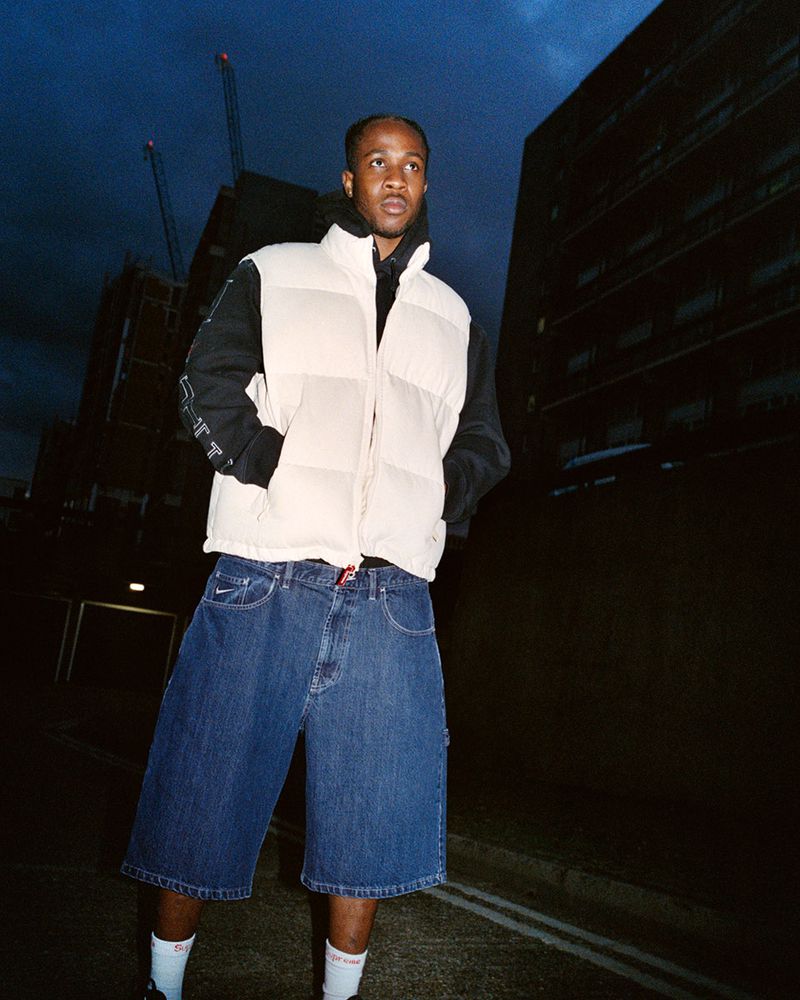 Adult male in a white vest and denim shorts standing on a city street in front of a tall building under a twilight sky.