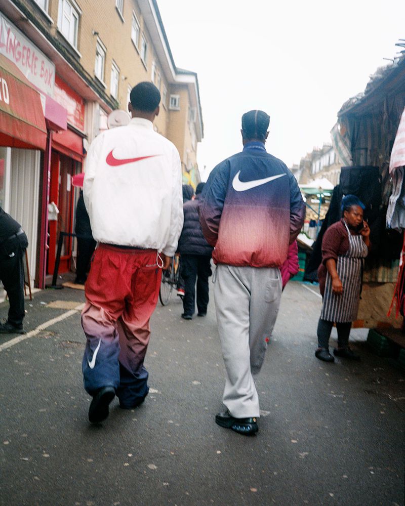 Two adults walk on a city street wearing matching Nike brand jackets and pants as vendors and buildings line the road.