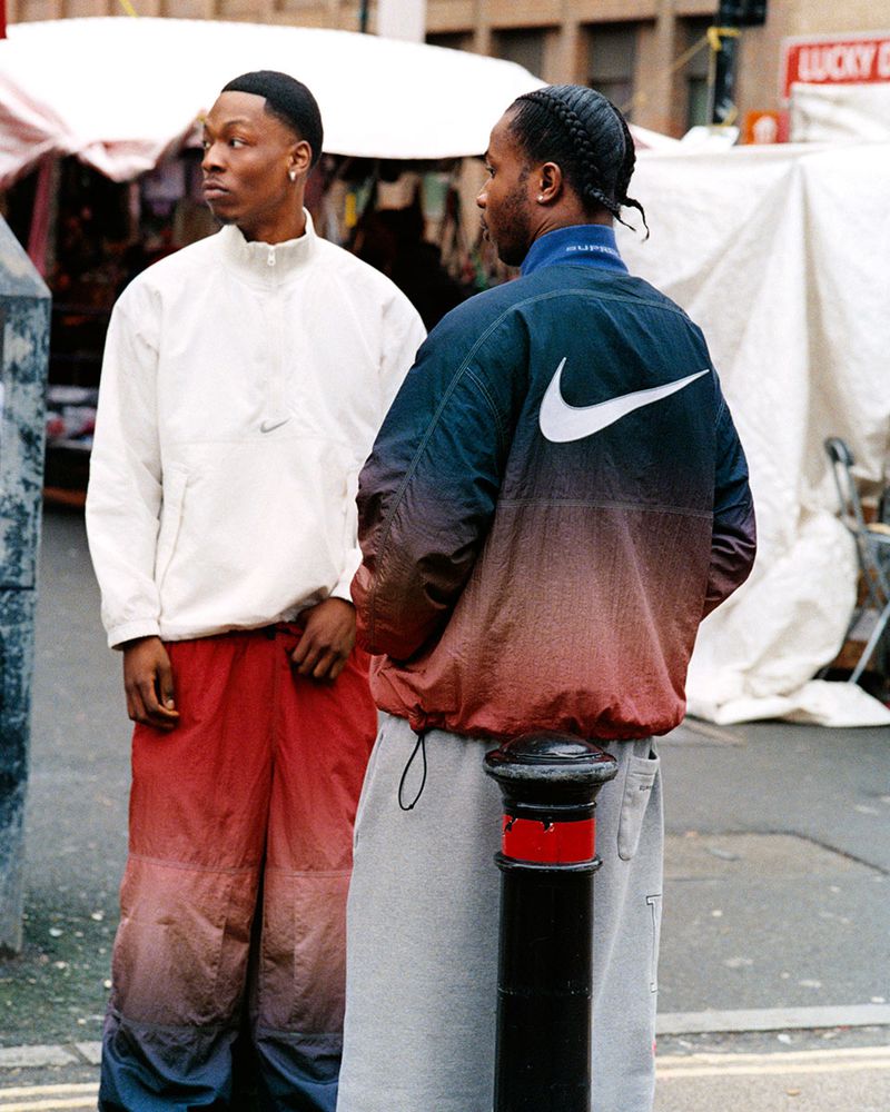 Two young adult men in streetwear stand outdoors on a city street. One wears a white jacket and the other wears a Nike jacket.
