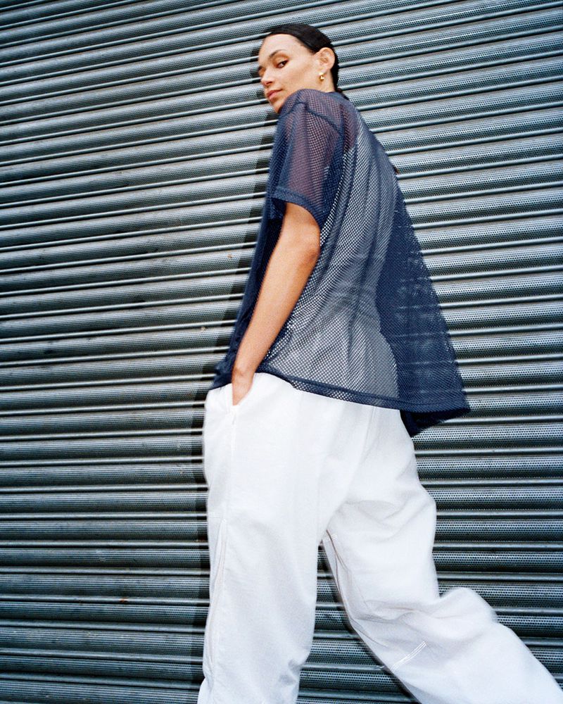 Adult female in a navy mesh shirt and white pants poses against a textured gray metal wall looking back at the camera.