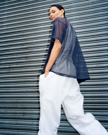 Adult female in a navy mesh shirt and white pants poses against a textured gray metal wall looking back at the camera.