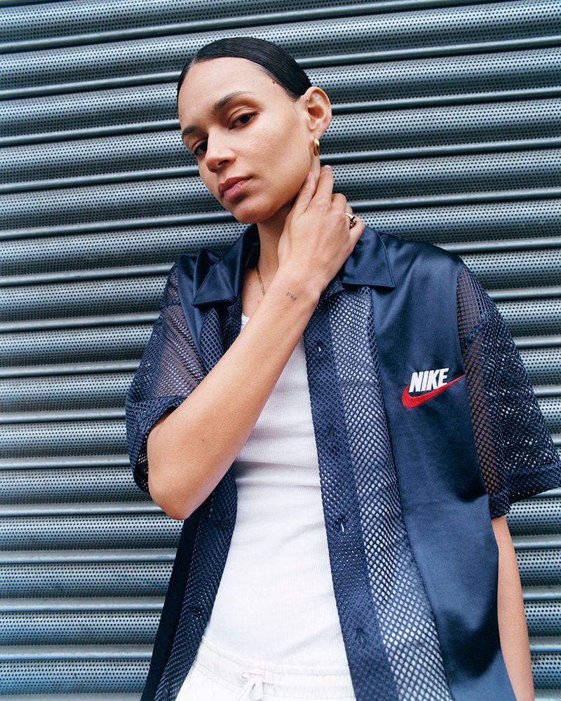 Woman in a navy mesh-paneled Nike top poses against a textured metal wall, showcasing fashion and athletic wear.