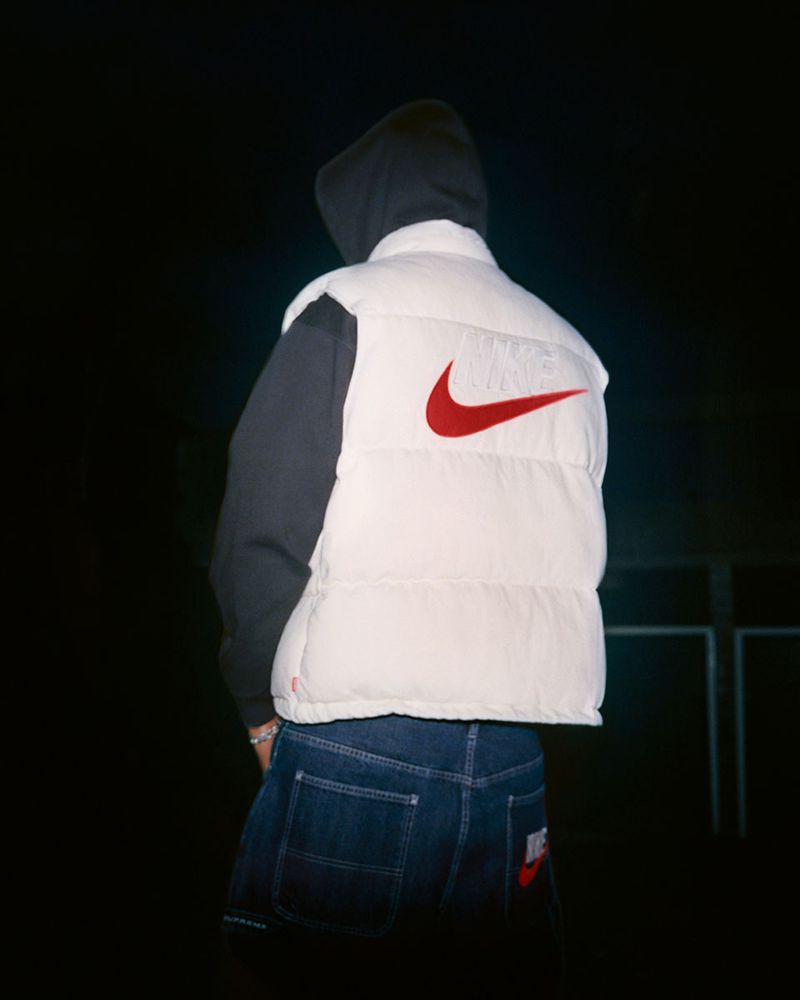 Person wearing a white Nike puffer vest and dark denim jeans, viewed from the back against a dark background.