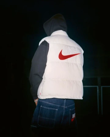 Person wearing a white Nike puffer vest and dark denim jeans, viewed from the back against a dark background.