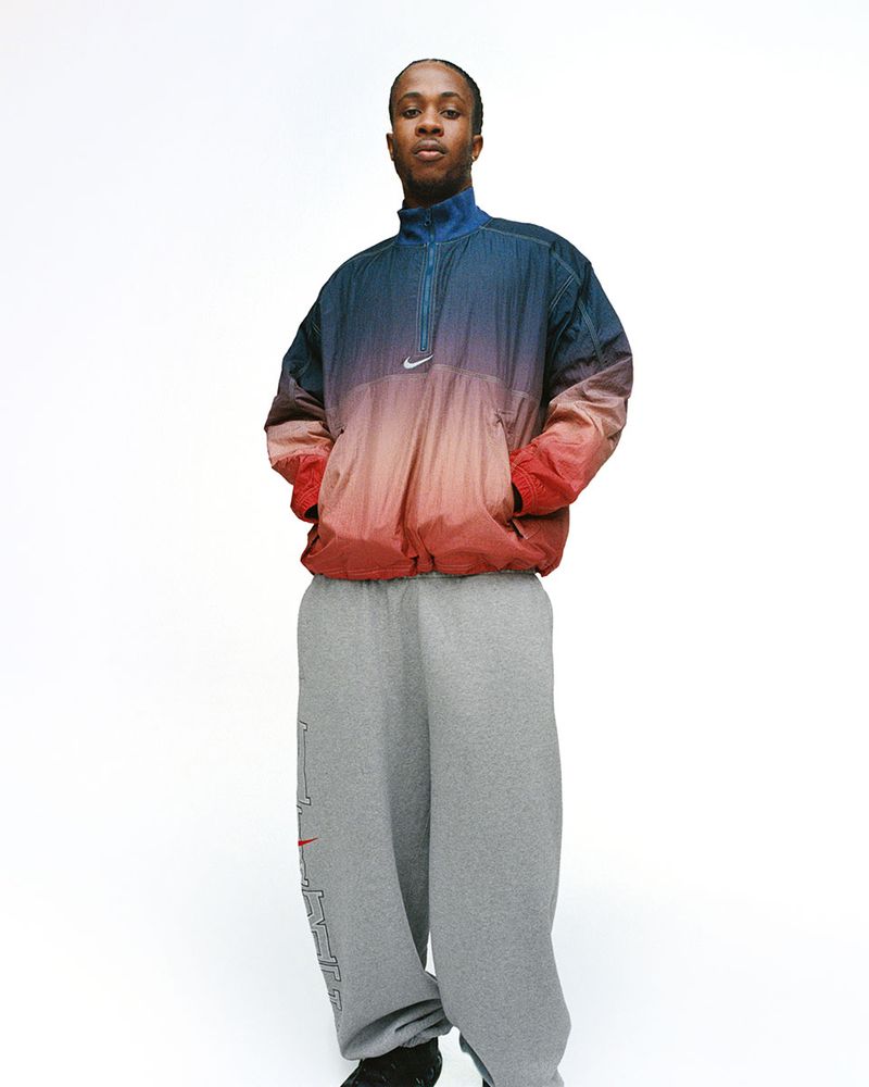 An adult male stands centered in a light studio wearing a blue-red gradient color top, gray sweatpants, and dark sneakers against a white backdrop.
