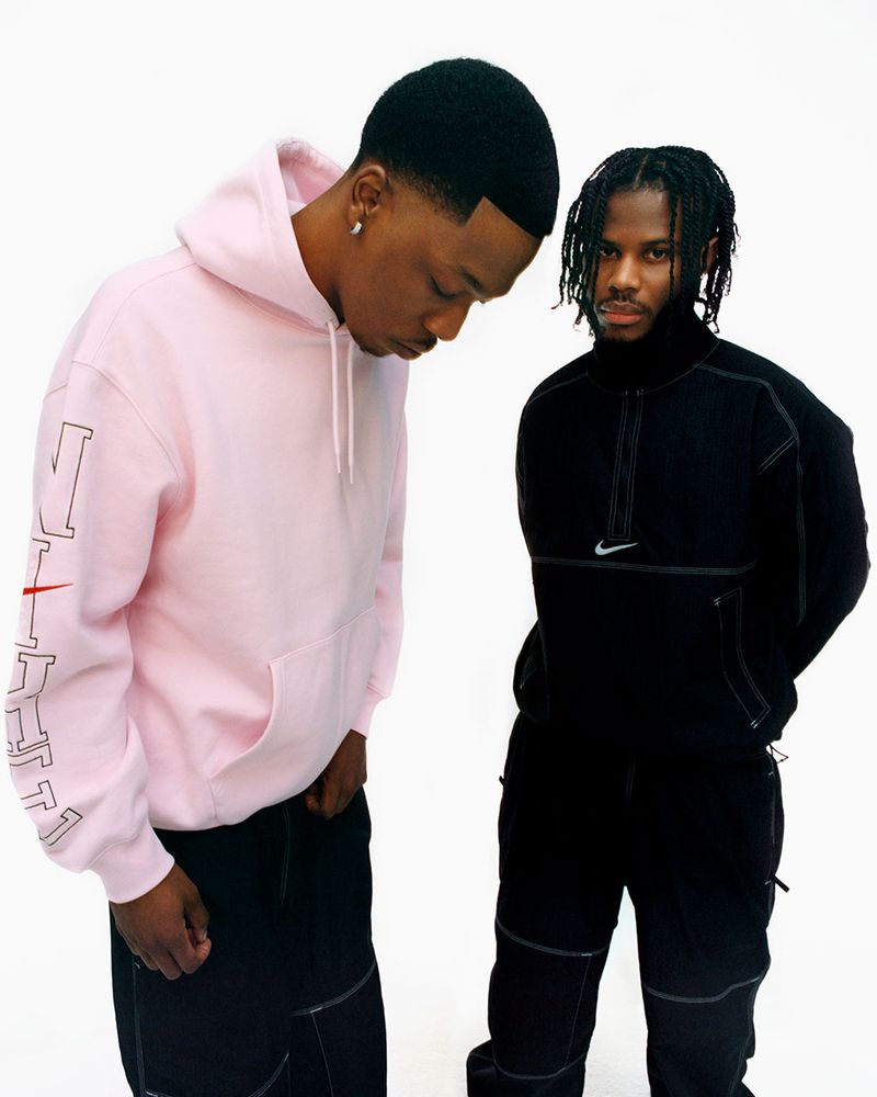 Two adult males pose in casual streetwear against a white backdrop, showcasing a pink hoodie and a black zip-up jacket with brand logos.