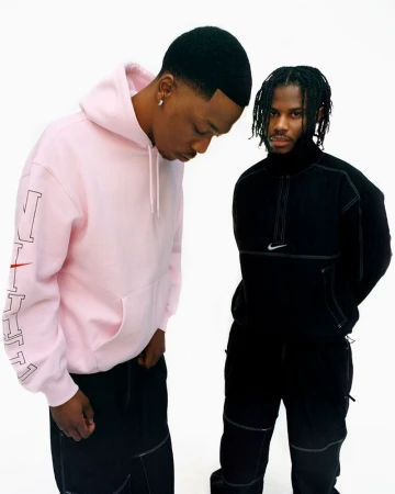 Two adult males pose in casual streetwear against a white backdrop, showcasing a pink hoodie and a black zip-up jacket with brand logos.