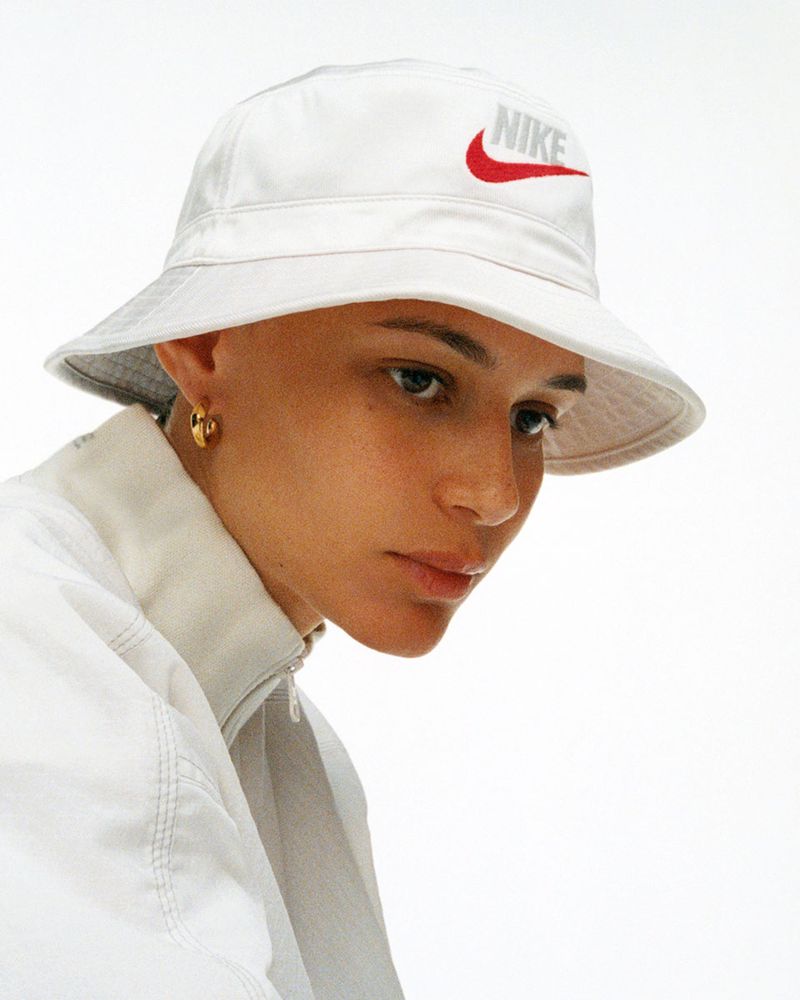 Young adult woman gazes downward wearing white Nike bucket hat and light jacket against a bright studio backdrop. Fashion shot.