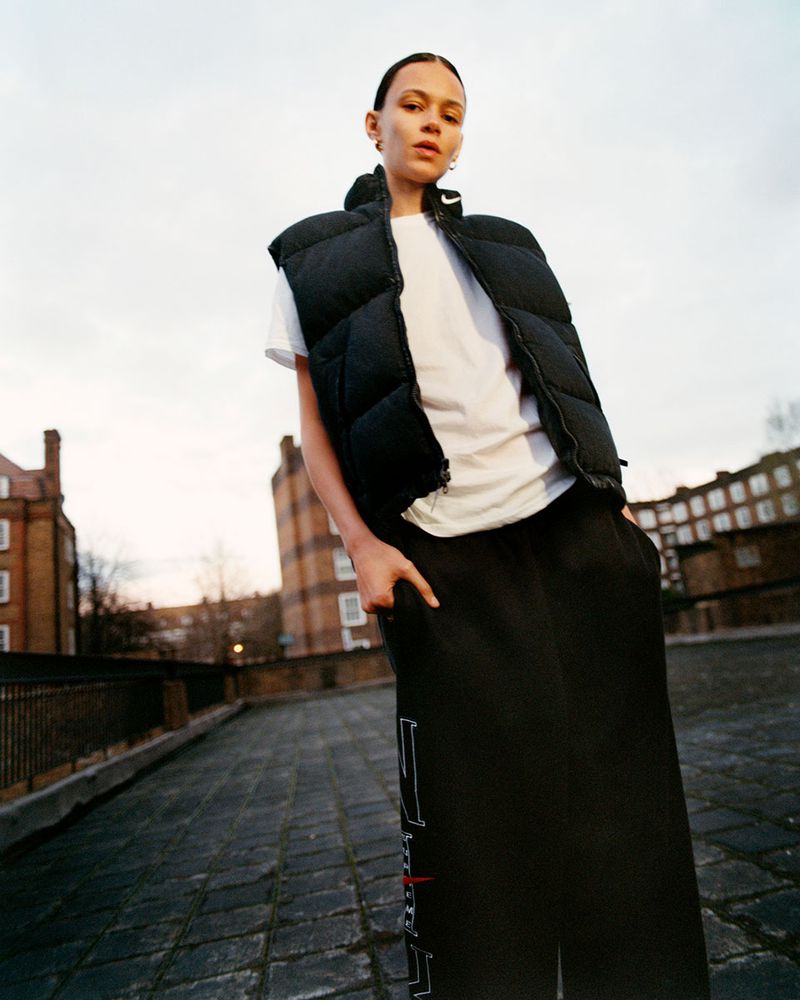 Woman in a dark puffy vest and black pants stands on a rooftop against a blurred city and sky backdrop in an eye-level medium shot.