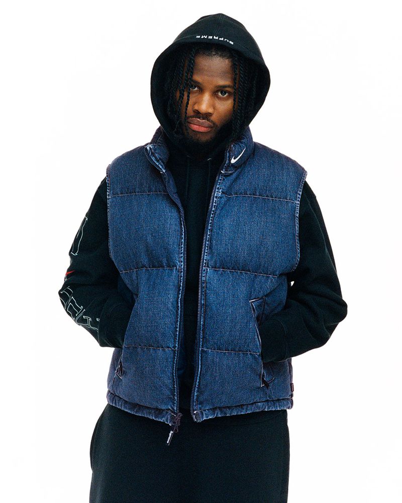 Adult male with dark hair wearing a denim puffer vest and a hooded sweatshirt against a plain white backdrop in a studio setting.