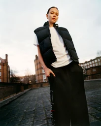 Model wearing a black puffer vest over a white t-shirt and dark pants on a paved rooftop at dusk