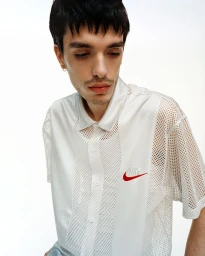Leo Bhanji wearing a white mesh short-sleeved collared shirt with a red Nike logo embroidery against a white studio background.