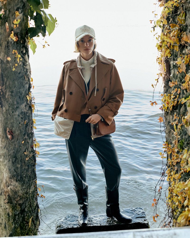 A young woman in a brown jacket, dark pants, and boots stands on a stone pier surrounded by water and trees, creating a fashion editorial.