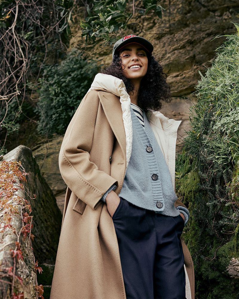 An adult woman is smiling in a tan overcoat, dark pants, and cap, posing on stone steps surrounded by leafy plants in an outdoor environment.