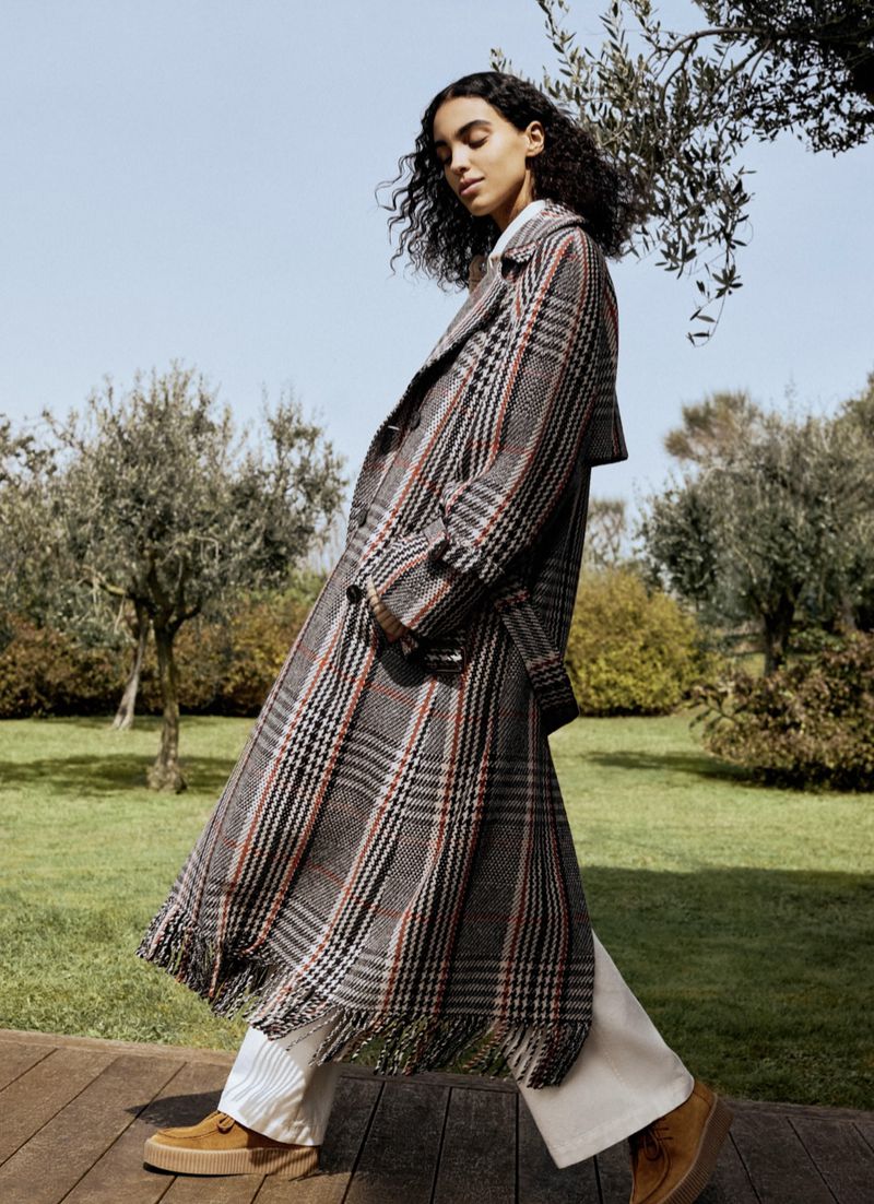 Model wearing a long plaid trench coat and white trousers walking in a green park