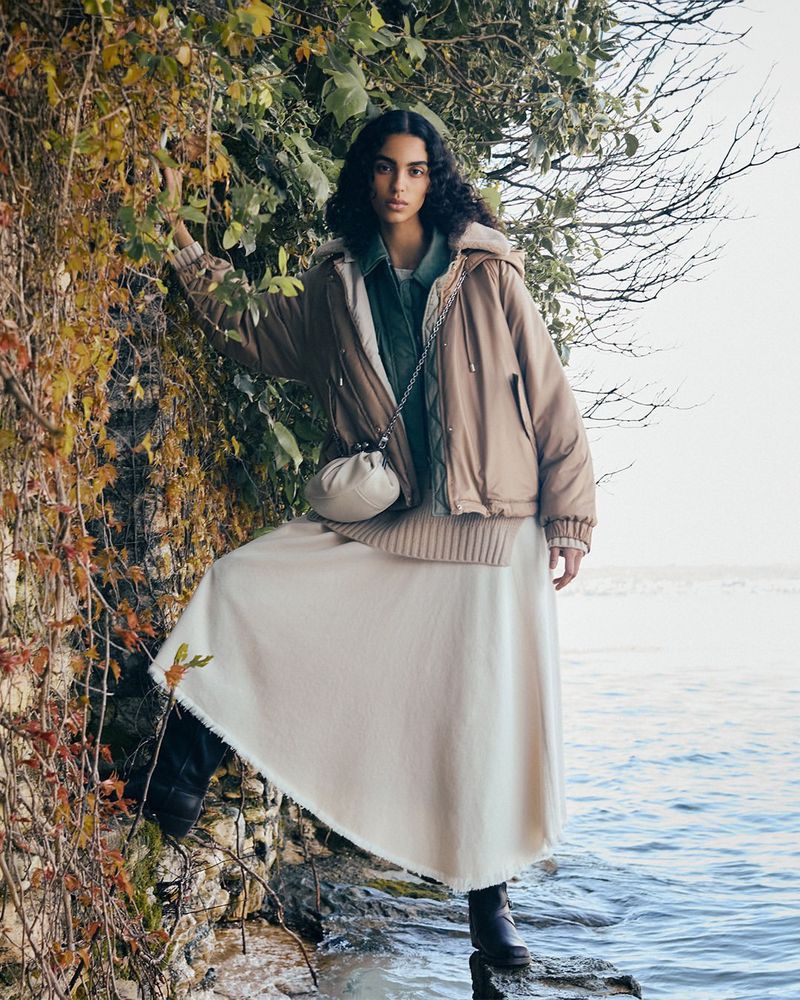A woman in a puffer jacket, long skirt, and boots poses outdoors near a stone wall and water, showcasing fashion and style.