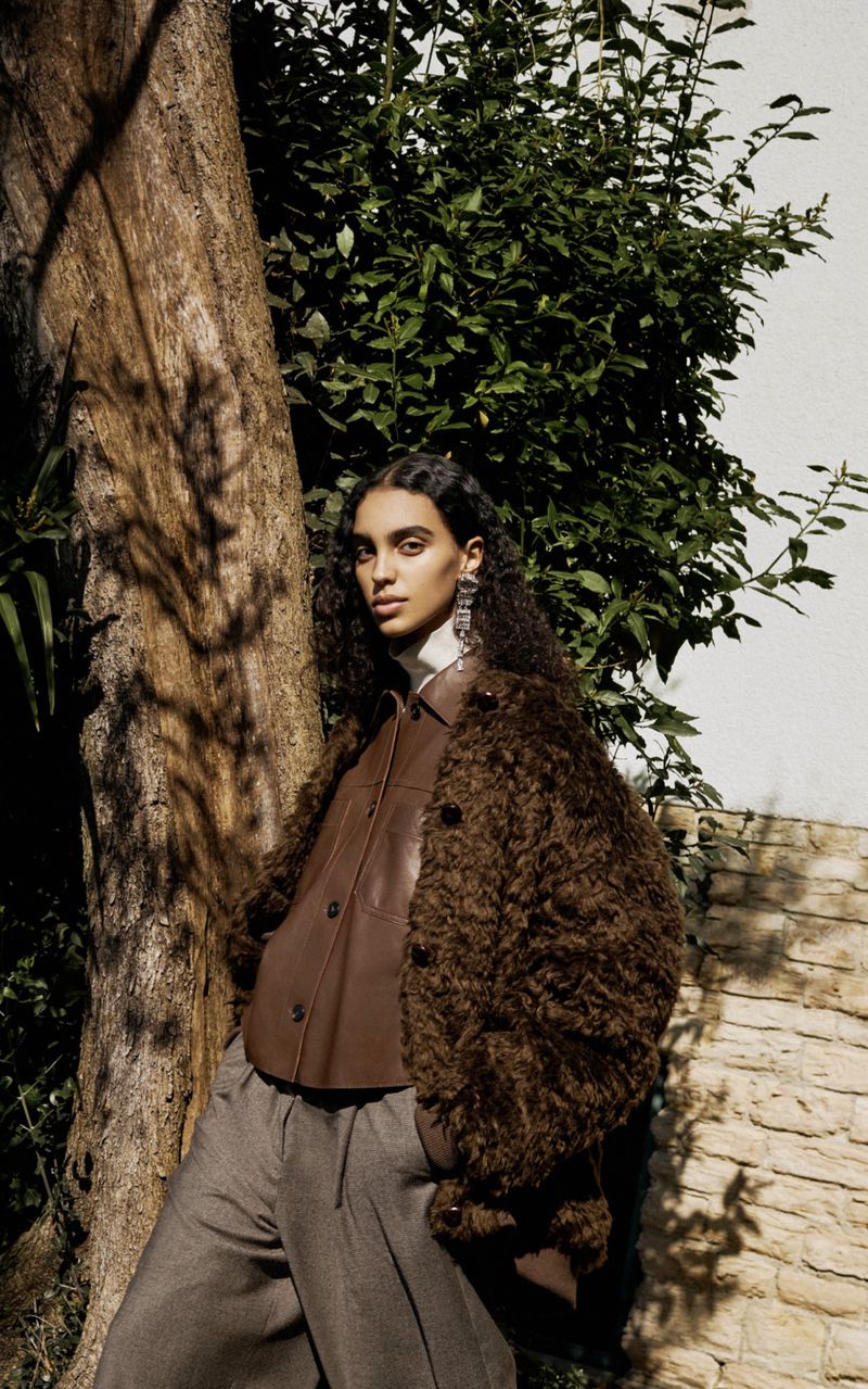 A curly-haired woman in a brown jacket and gray trousers poses outdoors by a tree, exuding a fashion-forward yet grounded aesthetic.