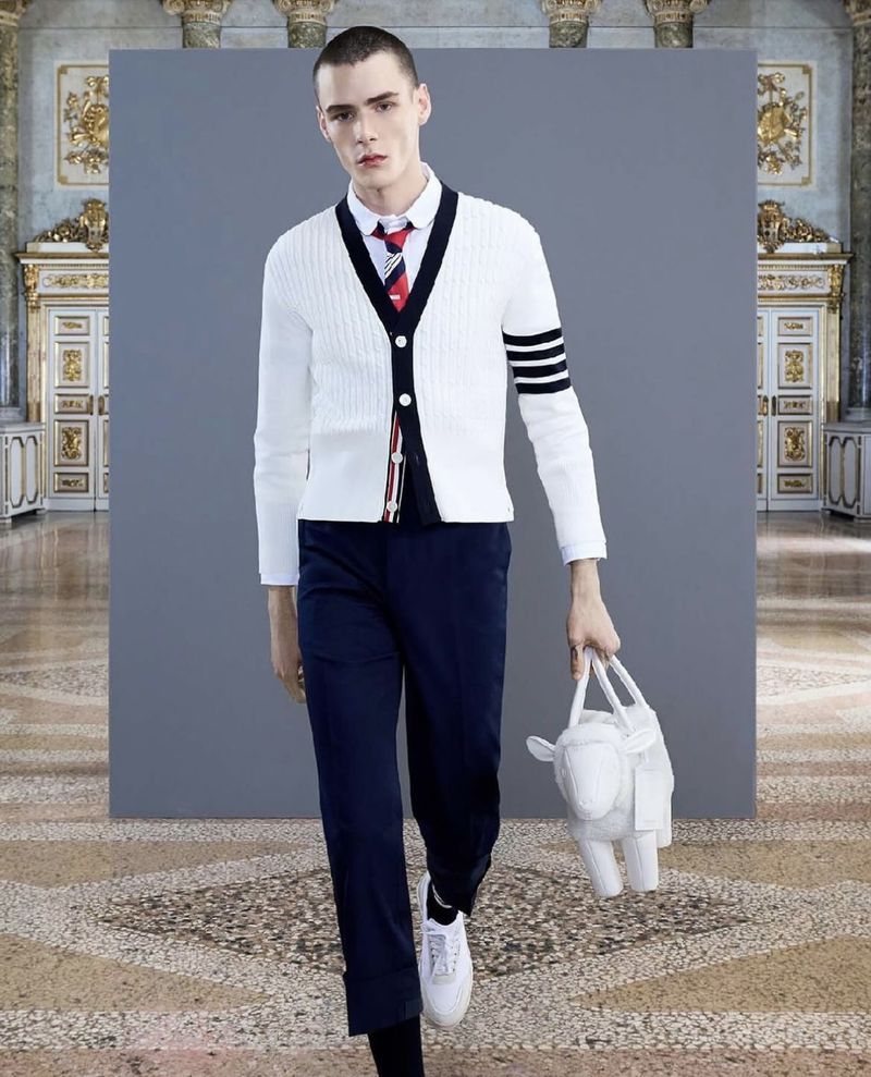Male fashion model in a white cable knit cardigan, dress shirt, tie, and blue trousers, holding a white sheep-shaped handbag.