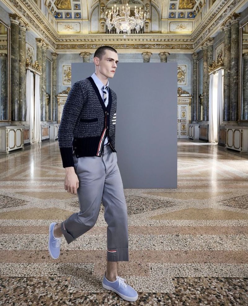 Fashion shot of a slender male model in a navy cardigan and grey pants walking in an ornate room with a patterned marble floor.