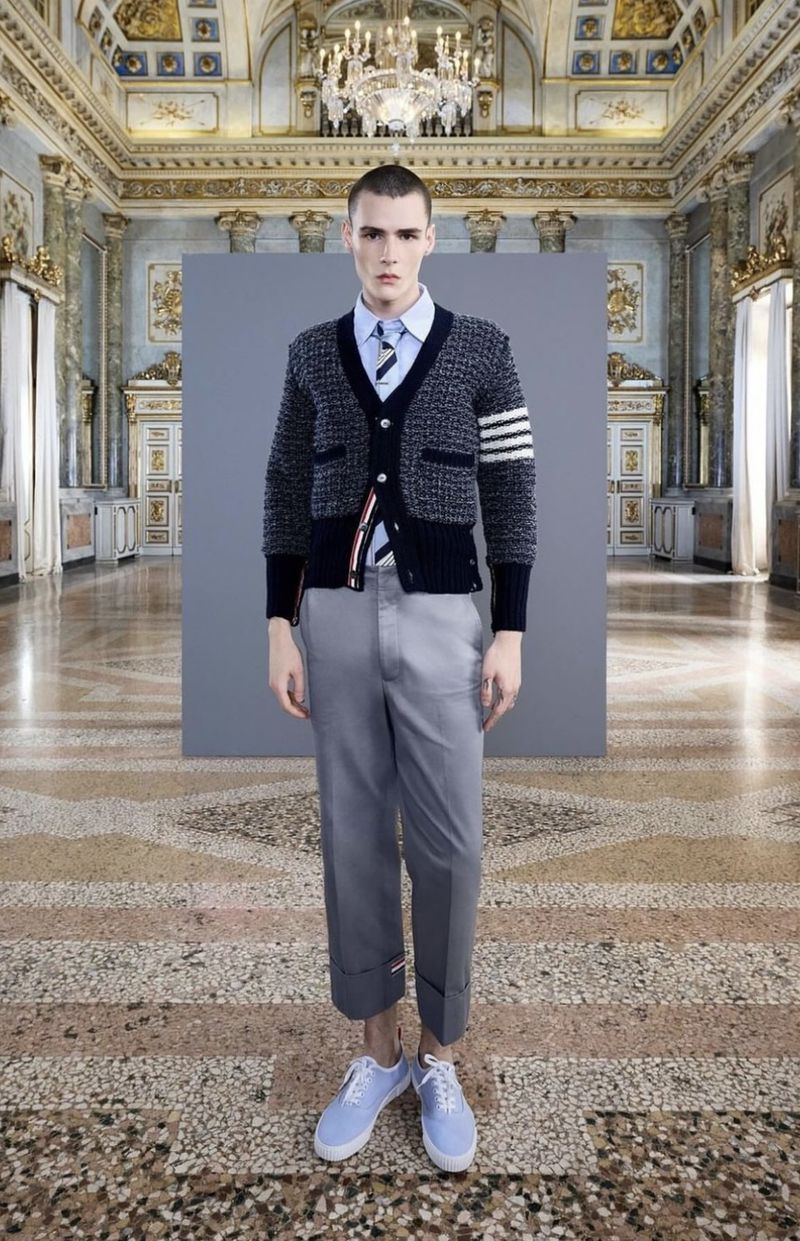 Male fashion model stands in ornate palace, wearing navy cardigan, light blue shirt, grey pants, light blue shoes. Formal indoor setting.