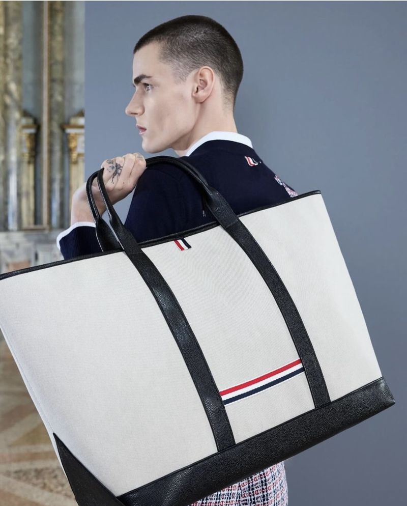 A stylish adult male with a short haircut is carrying a large white tote bag with black leather trim in an indoor setting.