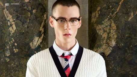 Model Paolo Grebic wearing optical glasses, a striped necktie, and a cream cable-knit sweater against a green marble background.