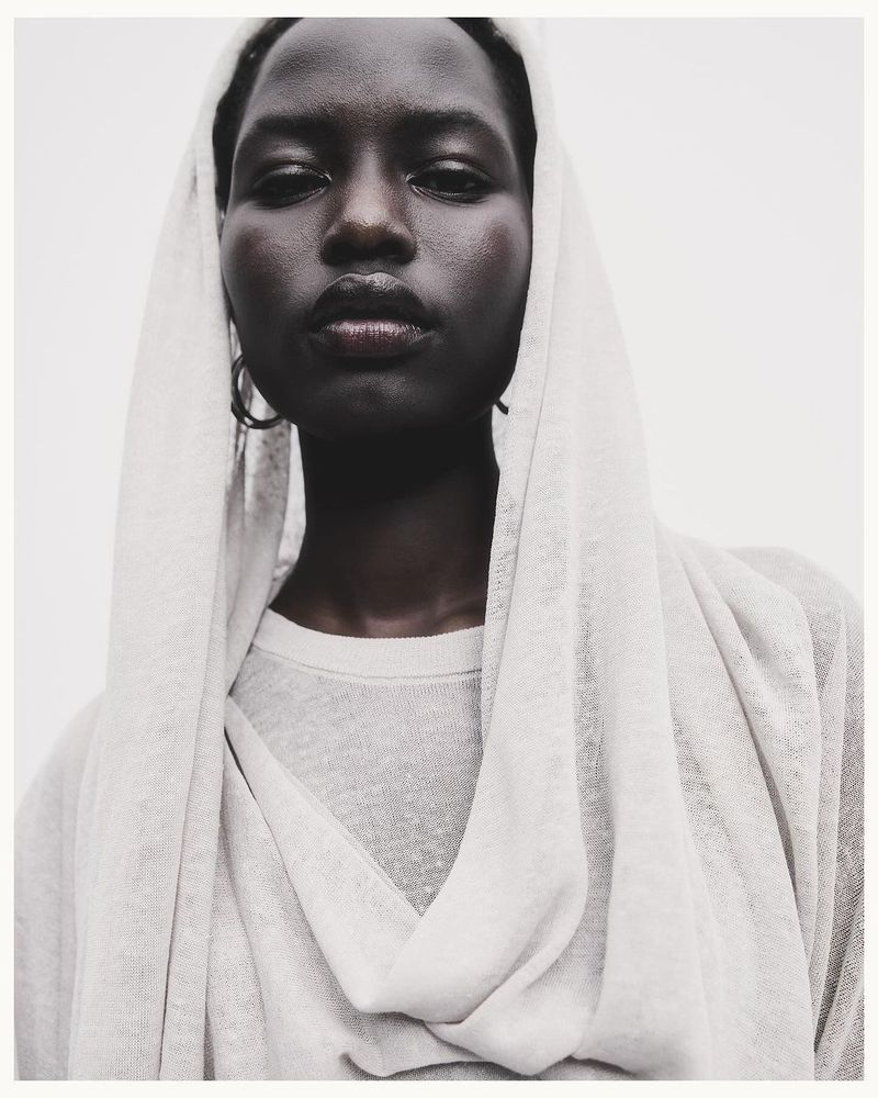 Close-up portrait of an African-descent adult female fashion model with dark hair, wearing a light gray top and head covering.