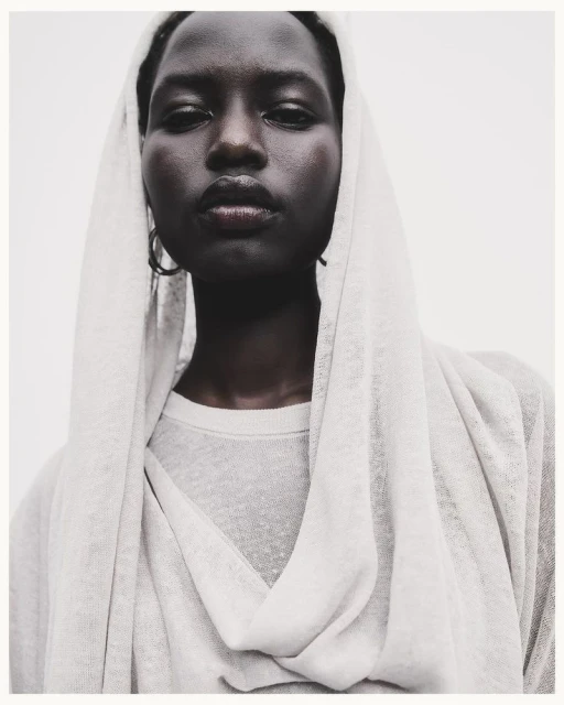 Close-up portrait of an African-descent adult female fashion model with dark hair, wearing a light gray top and head covering.
