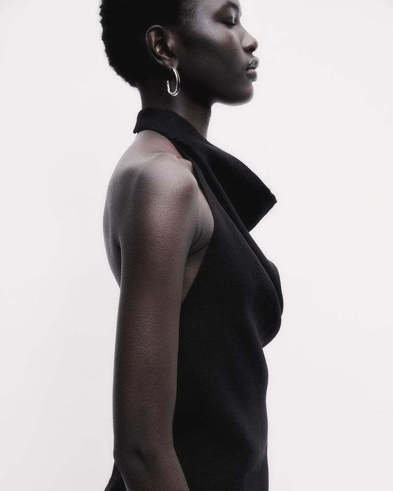 Profile of a Black woman with short hair wearing a black dress with a halter neckline against a solid white background in a studio shot.