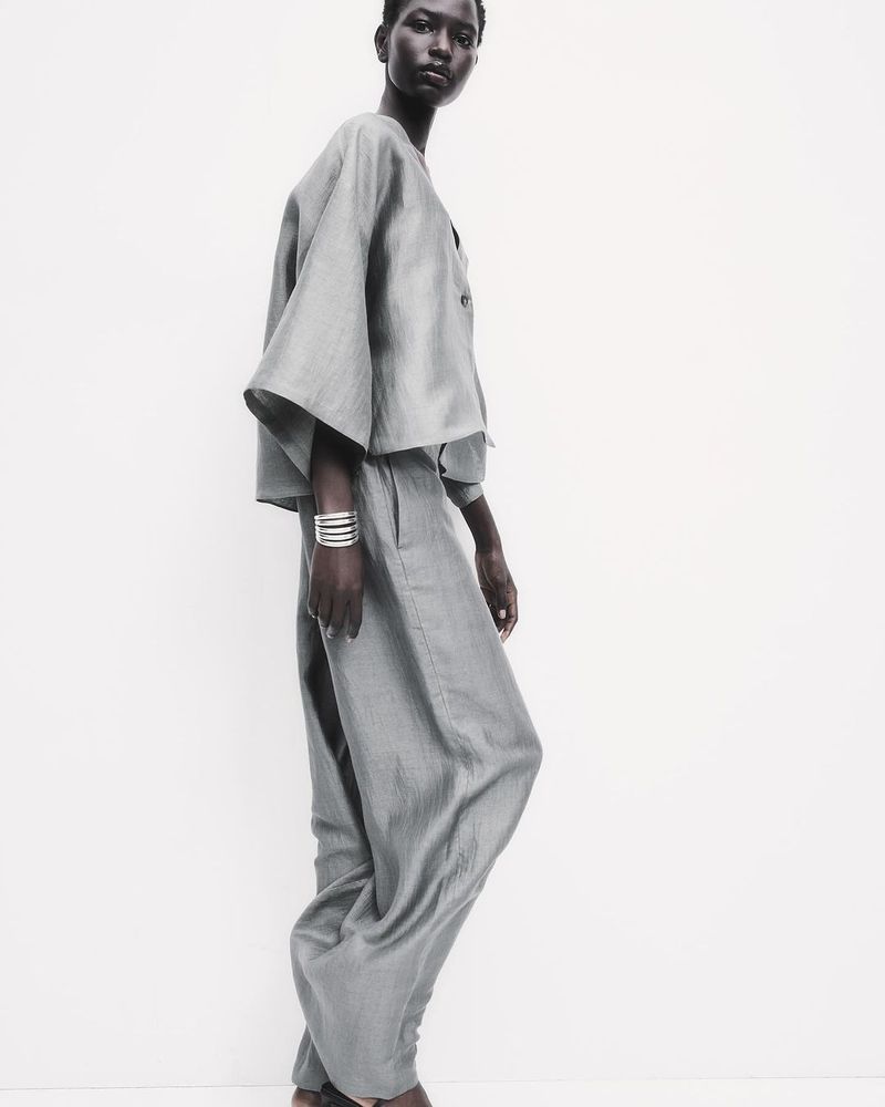 Fashion shot of an adult woman in loose fitting light grey two-piece outfit, accessorized with silver bracelets, posing indoors against white.