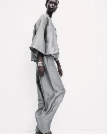 Fashion shot of an adult woman in loose fitting light grey two-piece outfit, accessorized with silver bracelets, posing indoors against white.