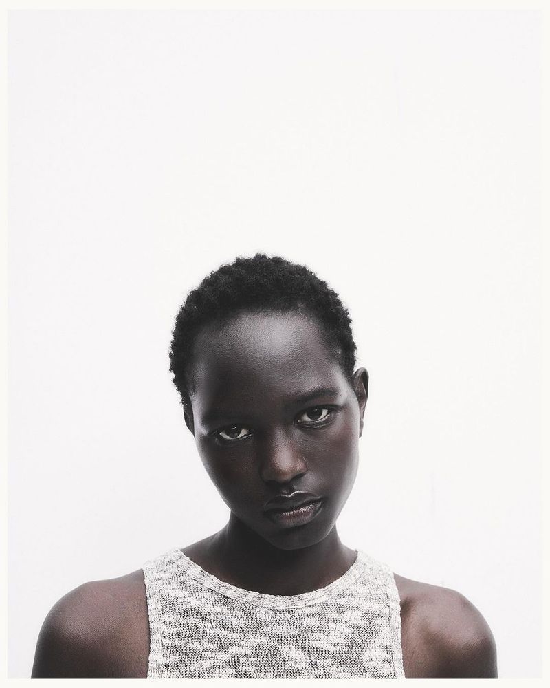 Portrait of a dark-skinned adult female model with short hair, wearing a light colored knit top, against a white backdrop in a photo studio.