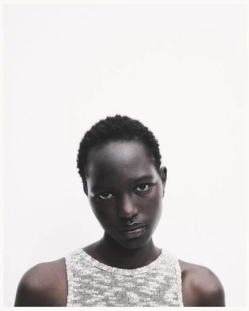 Portrait of a dark-skinned adult female model with short hair, wearing a light colored knit top, against a white backdrop in a photo studio.
