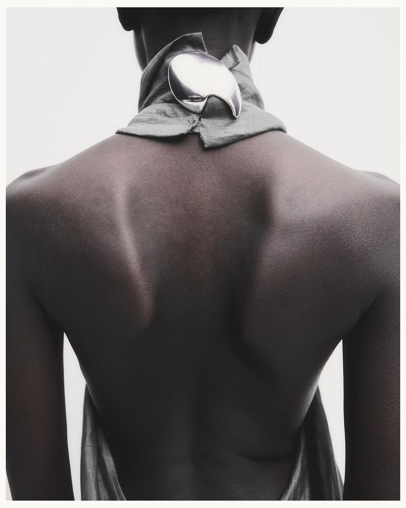 Back view of a person with dark skin wearing a light gray halter top, fastened with a large, abstract silver decorative clasp.