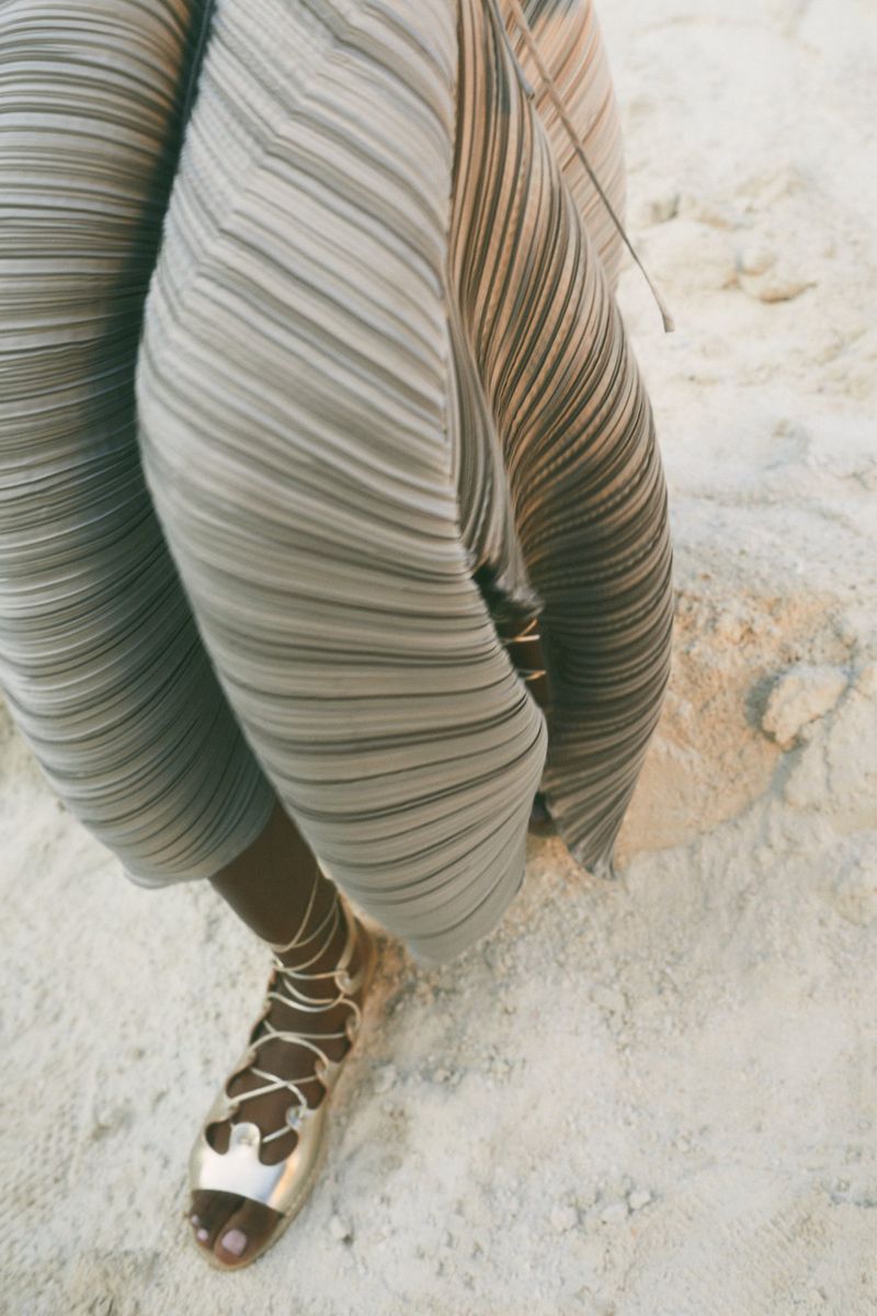 A model wears a pleated pants and metallic gladiator sandals while posing on a sandy surface, showcasing a fashion-forward outfit.