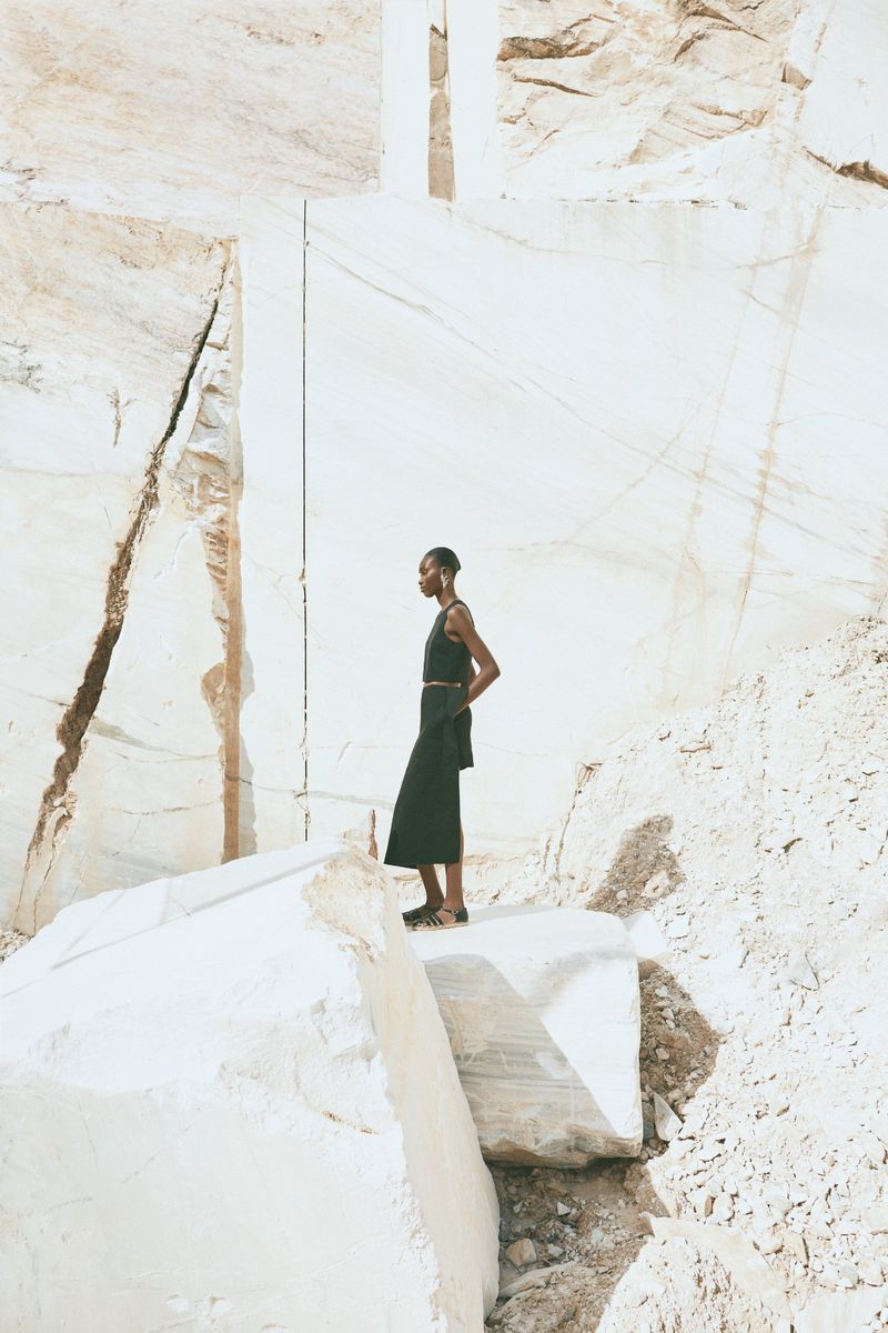 An adult female model stands on a stone block against a quarry wall, wearing a black top and skirt with a belt, set in a sunny outdoor setting.