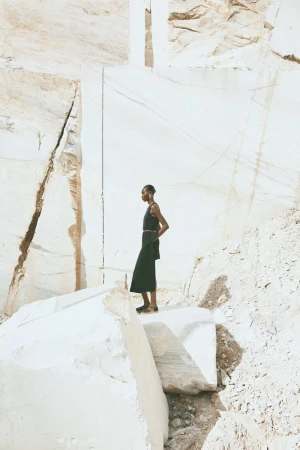 An adult female model stands on a stone block against a quarry wall, wearing a black top and skirt with a belt, set in a sunny outdoor setting.