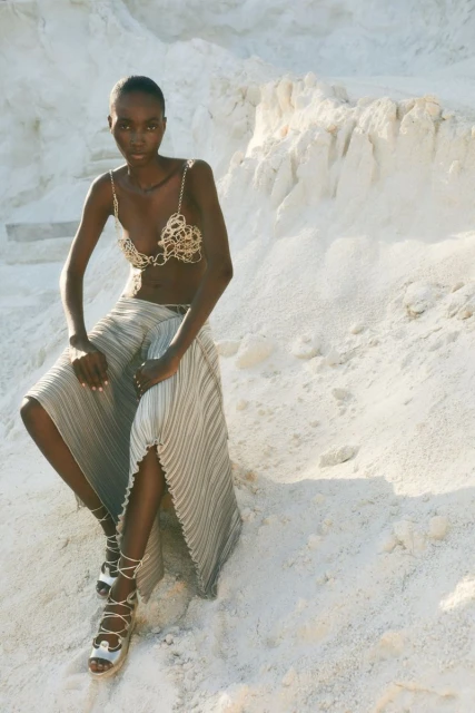 A slender Black woman poses in a decorative chain bra top and pleated skirt against a textured white background in a fashion-forward scene.