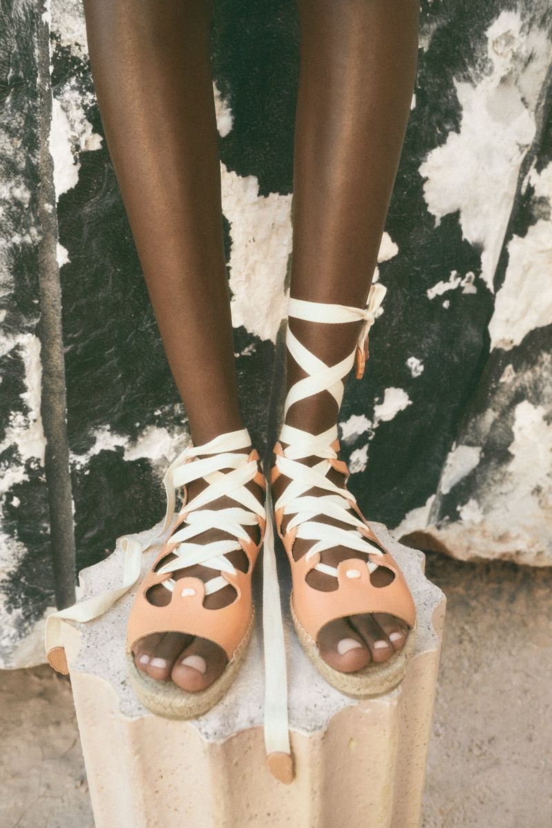 Close-up of a person with dark skin wearing strappy, peach-colored sandals with white ties on a grey stone stand.