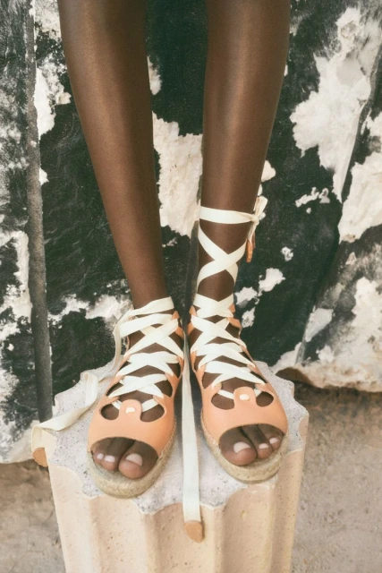 Close-up of a person with dark skin wearing strappy, peach-colored sandals with white ties on a grey stone stand.