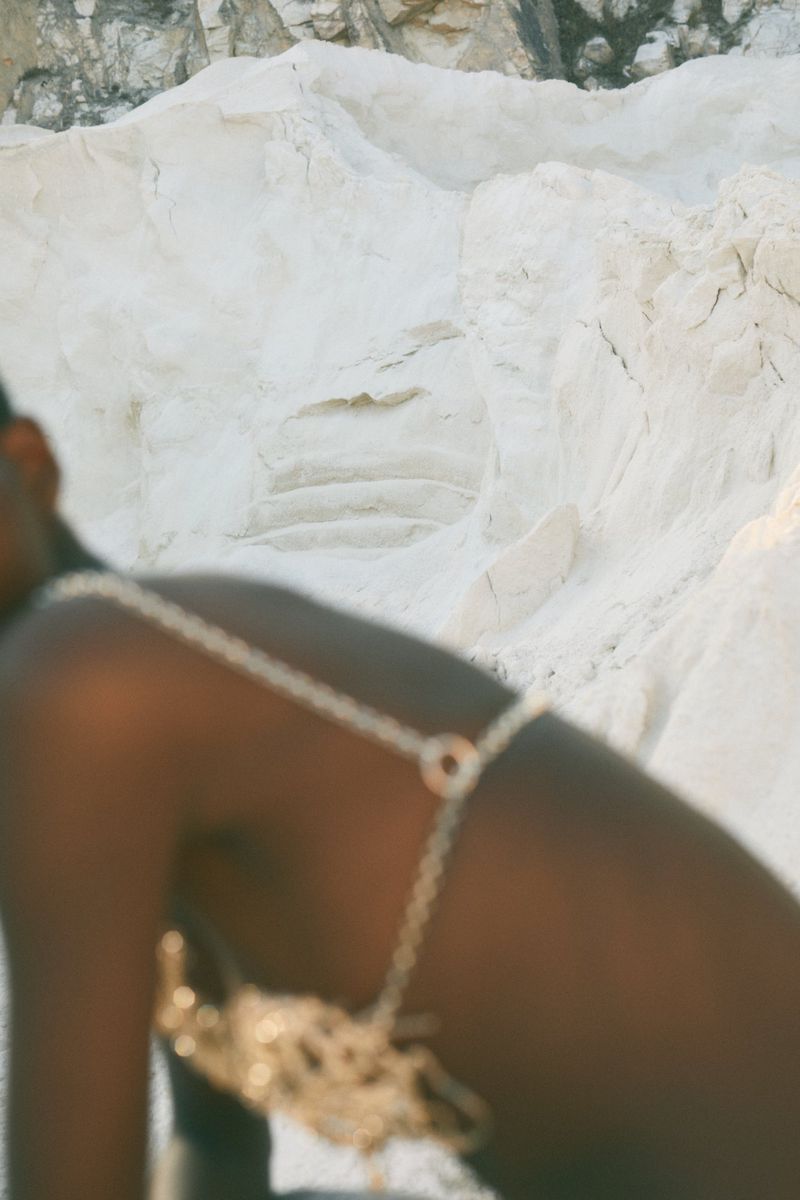 Woman in a gold chain bra stands out against a textured white stone background, creating a striking fashion statement.