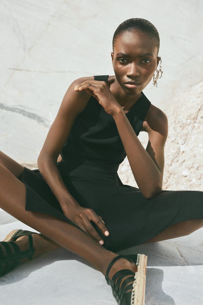 A dark-skinned woman in a black dress and strappy platform shoes poses cross-legged against a white mottled surface in an outdoor fashion shoot.