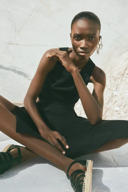 A dark-skinned woman in a black dress and strappy platform shoes poses cross-legged against a white mottled surface in an outdoor fashion shoot.